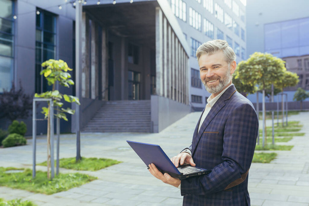 Portrait of mature businessman outside modern office building, senior gray-haired investor with laptop smiling and looking at camera, successful man in business suit holding netbook. - Φωτογραφία, εικόνα