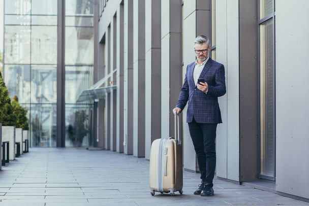 Business trip, senior gray-haired businessman outside hotel using phone, mature boss with big suitcase, investor in business suit and glasses. - Foto, afbeelding
