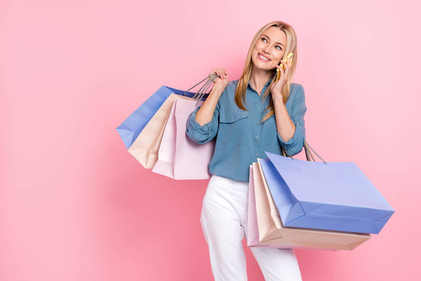 Photo of positive cheerful woman wear blue blouse talk on phone hold shopping bags look empty space isolated on pink color background. - 写真・画像