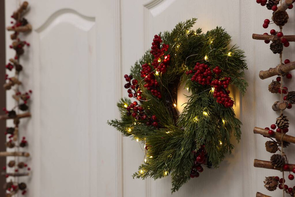 Beautiful Christmas wreath with red berries and fairy lights hanging on white door, space for text - Photo, Image