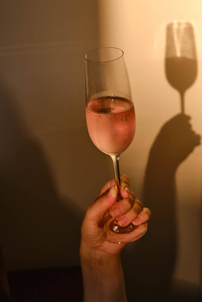 Glass of rose wine in the hands of a girl in the rays of the setting sun at home. Tasting of alcoholic beverages. Celebrate and enjoy the moment. Romantic evening aperitif. Close-up glass of wine - 写真・画像
