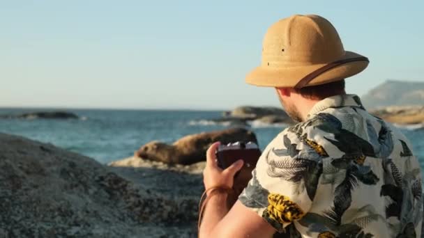 Ein männlicher Reisender mit Safari-Hut steht neben einer jungen Robbe auf dem Ozean. Ein männlicher Fotograf fotografiert am Strand eine schlafende Robbe in der Sonne. Soloreise durch den Amazonas - Filmmaterial, Video