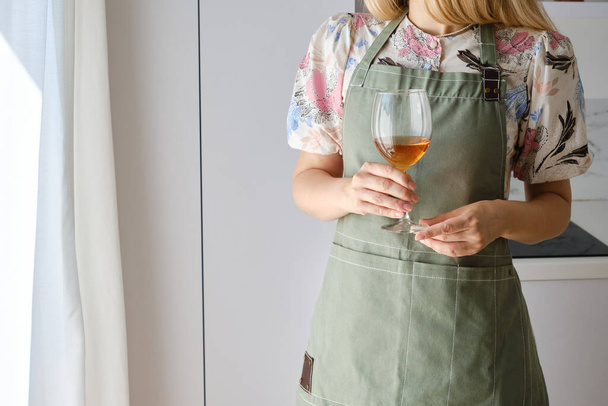 A woman in a kitchen apron and with a glass of white wine in a modern kitchen. Wine tasting. Cooking at home in uniform, protection apparel. Green fabric apron, casual wear - Foto, imagen