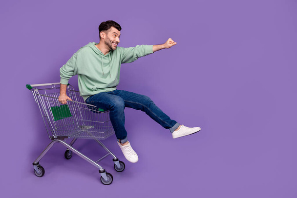 Full body photo of overjoyed cheerful person sit ride market trolley look empty space isolated on violet color background. - Photo, image