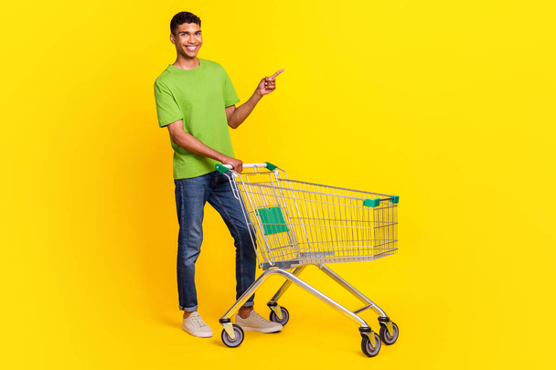 Full size photo of positive man market trolley indicate finger empty space isolated on yellow color background. - Zdjęcie, obraz