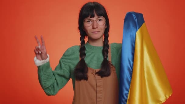 Mladá nerd žena s copánky v brýlích drží Ukrajinu národní vlajku, ukazuje vítězství znamení proti válce, doufat v úspěch a vyhrát. Dívka dělá poklidné gesto, usmívá se s laskavým optimistickým výrazem - Záběry, video
