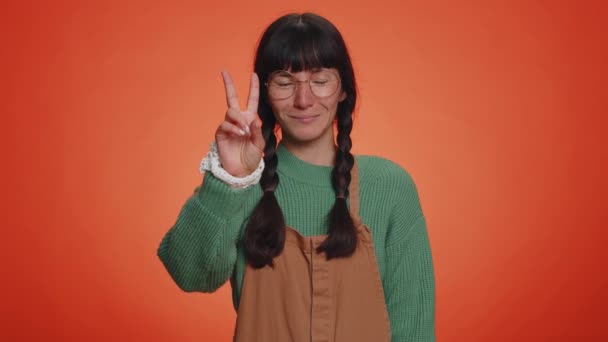 Hipster woman in green sweater showing victory sign, hoping for success and win, doing peace gesture, smiling with kind optimistic expression. Young adult girl isolated on orange studio background - Footage, Video
