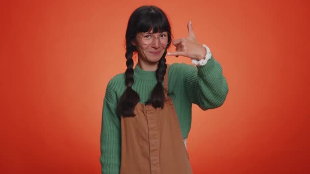Rufen Sie mich an, hier ist die Kontaktnummer. Junge Nerd-Frau mit Zöpfen in Brille, die in die Kamera schaut und Handygesten macht wie "Hallo, du rufst mich zurück, Gespräch". Latin Mädchen isoliert auf orangefarbenem Hintergrund - Filmmaterial, Video