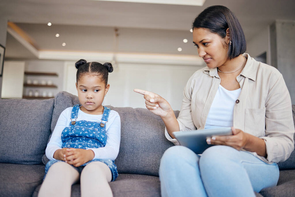 Mixed race woman pointing to scold and discipline her little daughter about internet use on digital tablet at home. naughty girl looking sad while mother punishes her for playing too many games onlin. - Zdjęcie, obraz