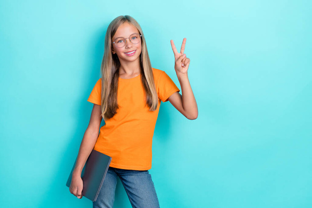 Photo of smart cute little girl wear orange t-shirt toothy smile hold laptop v-sign toothy smile enjoy home education isolated on aquamarine color background. - Fotó, kép