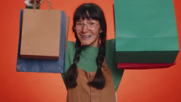 Χαρούμενη τουρίστρια γυναίκα σε πράσινο πουλόβερ δείχνει τσάντες ψώνια, διαφημίσεις εκπτώσεις, χαμογελώντας αναζητούν έκπληκτος με χαμηλές τιμές, ψώνια στις διακοπές Black Friday. Νεαρό κορίτσι χιλιετηρίδας σε πορτοκαλί φόντο - Πλάνα, βίντεο