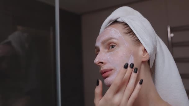 Donna con asciugamano avvolto intorno alla testa applicando maschera viso argilla prima specchio da bagno. routine serale quotidiana pulizia del viso, cura della pelle, peeling, idratante e concetto di trattamento di bellezza - Filmati, video