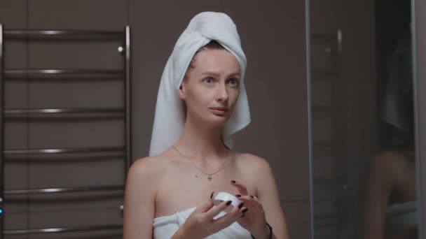 Mujer con toalla envuelta alrededor de su cabeza aplicando crema hidratante antes del espejo de baño. Rutina nocturna diaria - limpieza facial, cuidado de la piel, pelado concepto de tratamiento de belleza - Imágenes, Vídeo