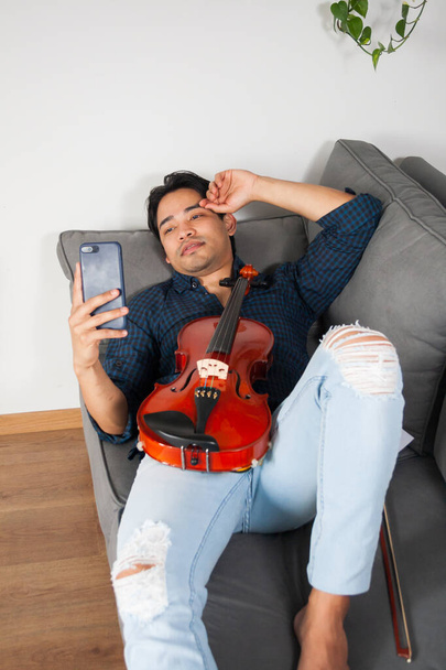 Young Hispanic male musician, smiling on a cell phone video call, lying on his couch at home. - Foto, Bild