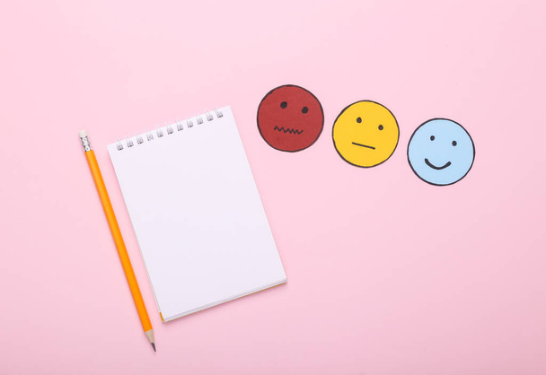 Concept d'enquête de satisfaction client. Carnet avec icône de papier visage heureux, neutre et triste sur fond rose. Vue du dessus - Photo, image