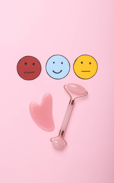Beauty-Branchenbericht. Umfrage zur Kundenzufriedenheit. Jade-Gesichtsmassagerolle mit fröhlichem, neutralem und traurigem Gesichtspapier-Symbol auf rosa Hintergrund. Ansicht von oben - Foto, Bild