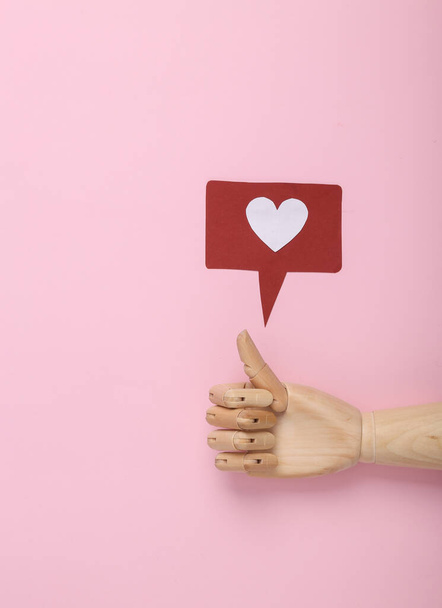 Mão de madeira com polegares para cima e um ícone de notificação semelhante em um fundo rosa. Redes sociais. Avaliação, feedback dos seguidores - Foto, Imagem