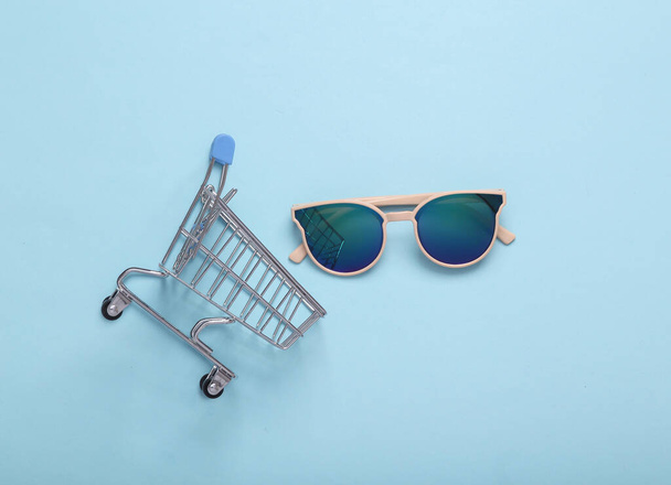 Concetto di shopping. Carrello shopping con occhiali da sole su sfondo blu pastello. posa piatta - Foto, immagini