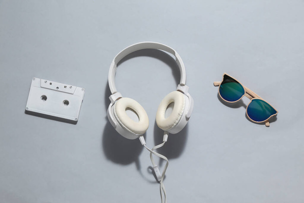 Creativo bodegón mínimo, auriculares estéreo con gafas de sol y cassette de audio sobre fondo gris brillante. Jóvenes, accesorios hipster, retro 80s. Vista superior - Foto, Imagen