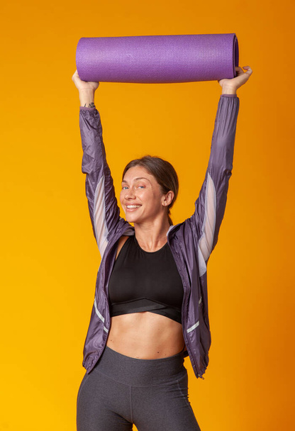 Cool cheerful athletic woman with yoga mat over her head on a yellow background. Healthy lifestyle - Photo, Image