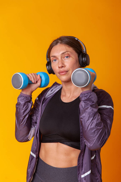 Workout concept. Happy athletic woman in sportswear posing with dumbbells in her hands on yellow background. Bodybuilding and fitness. Healthy lifestyle - Photo, Image