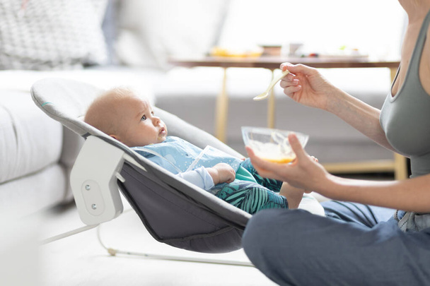 Mother spoon feeding her baby boy child in baby chair with fruit puree. Baby solid food introduction concept - Photo, image