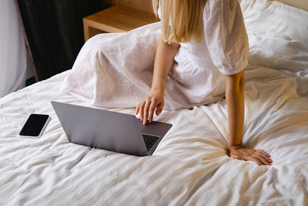 Girl works with a laptop and a smartphone in bed at home or in hotels. Distant work. Office at home. Woman in the bedroom is typing on a laptop, distance learning for students, surfing the Internet - Фото, изображение
