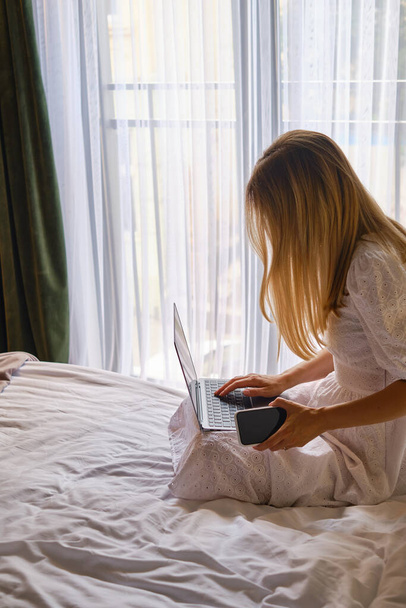 Girl works with a laptop and a smartphone in bed at home or in hotels. Distant work. Office at home. Woman in the bedroom is typing on a laptop, distance learning for students, surfing the Internet - Φωτογραφία, εικόνα