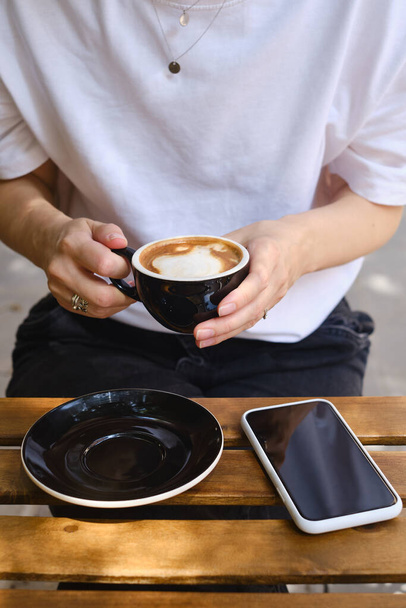 Girl drinks coffee from a black cup and uses a smartphone. A woman is holding a mobile phone, sending a text message or using an application on her mobile phone. Drink cappuccino or latte in a cafe - Φωτογραφία, εικόνα