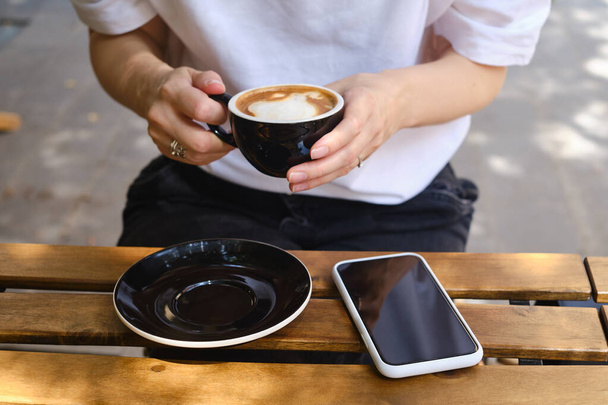 Girl drinks coffee from a black cup and uses a smartphone. A woman is holding a mobile phone, sending a text message or using an application on her mobile phone. Drink cappuccino or latte in a cafe - Photo, Image