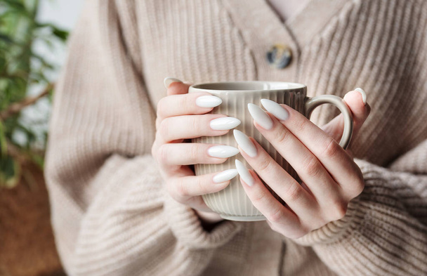 Hermosas manos de una joven con manicura blanca en las uñas. Chica en un suéter sosteniendo una taza de té - Foto, imagen