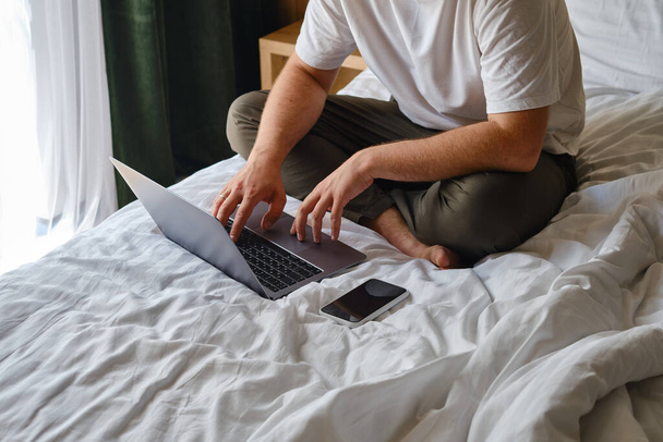 A man works with a laptop and smartphone in bed at home or in hotels. Remote work. Office at home. A man in the bedroom is typing on a laptop, distance learning for students, surfing the Internet - Foto, Imagem