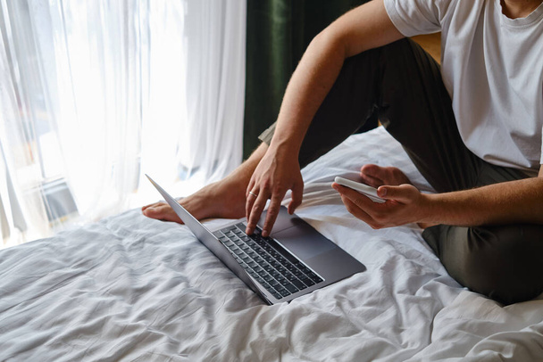 A man works with a laptop and smartphone in bed at home or in hotels. Remote work. Office at home. A man in the bedroom is typing on a laptop, distance learning for students, surfing the Internet - Photo, Image