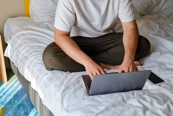 A man works with a laptop and smartphone in bed at home or in hotels. Remote work. Office at home. A man in the bedroom is typing on a laptop, distance learning for students, surfing the Internet - Photo, Image