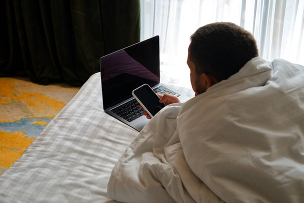 A man works with a laptop and smartphone in bed at home or in hotels. Remote work. Office at home. A man in the bedroom is typing on a laptop, distance learning for students, surfing the Internet - Фото, зображення