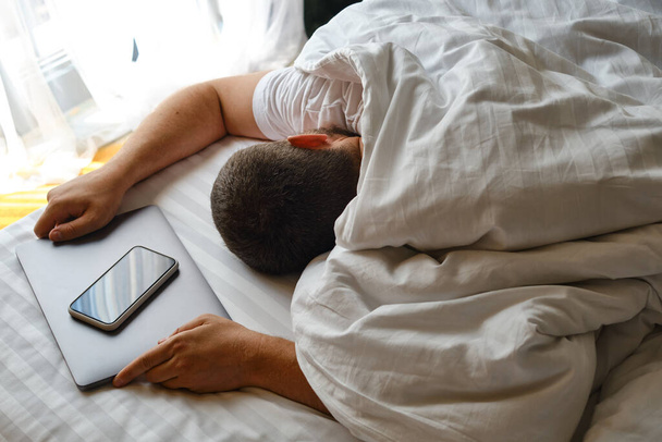 The man fell asleep in bed with a laptop and a telephone. Man working on laptop and phone in bed. Freelancer home office. Gadget addiction. Relax after work day concept - Zdjęcie, obraz
