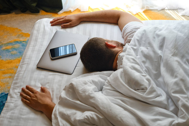 The man fell asleep in bed with a laptop and a telephone. Man working on laptop and phone in bed. Freelancer home office. Gadget addiction. Relax after work day concept - Foto, afbeelding