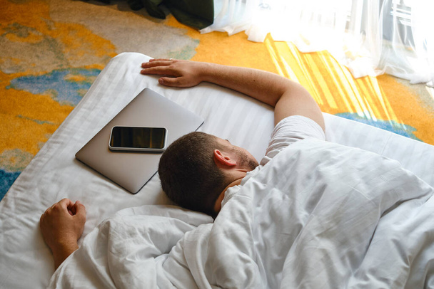 The man fell asleep in bed with a laptop and a telephone. Man working on laptop and phone in bed. Freelancer home office. Gadget addiction. Relax after work day concept - Фото, изображение