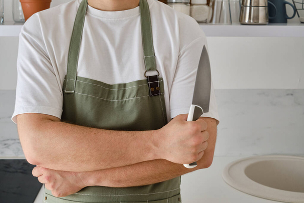 A man in a kitchen apron and a knife stands in a modern kitchen. Cooking at home in uniform, protection apparel. Green fabric apron, casual wear. A man in an apron prepares to prepare a meal - Фото, зображення