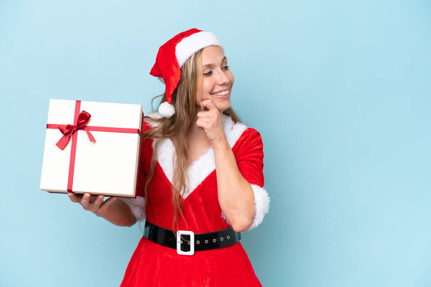 Young blonde woman dressed as Mama claus holding a present isolated on blue background thinking an idea and looking side - Photo, Image