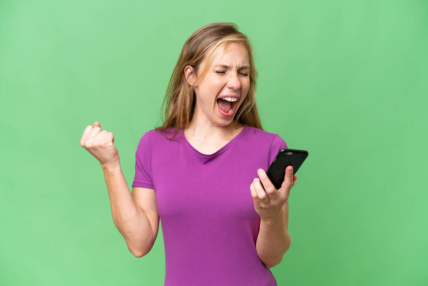 Молодая блондинка на изолированном фоне с телефоном в победной позиции - Фото, изображение