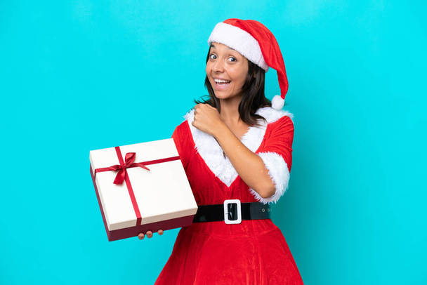 Anne Noel gibi giyinmiş genç İspanyol kadın mavi arka planda izole edilmiş bir hediyeyle zaferi kutluyor. - Fotoğraf, Görsel