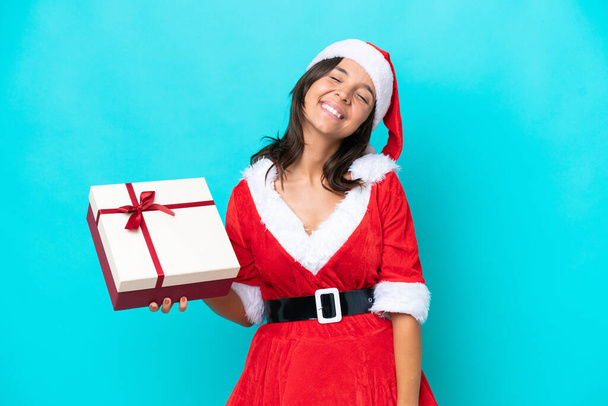 Anne Noel gibi giyinmiş genç İspanyol kadın elinde mavi arka planda izole edilmiş bir hediyeyle gülüyor. - Fotoğraf, Görsel