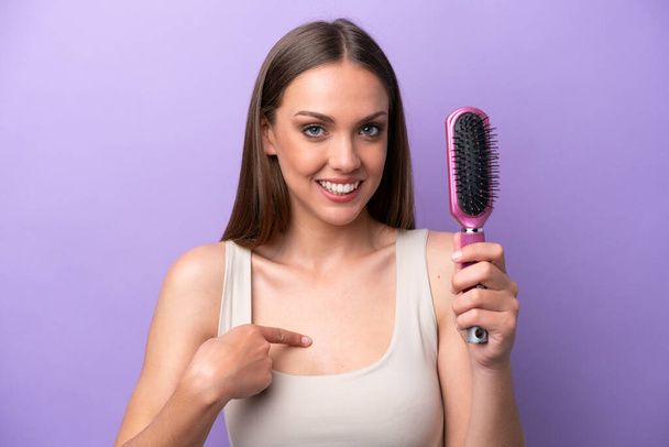 Jovem caucasiana segurando escova de cabelo isolada em fundo roxo com expressão facial surpresa - Foto, Imagem