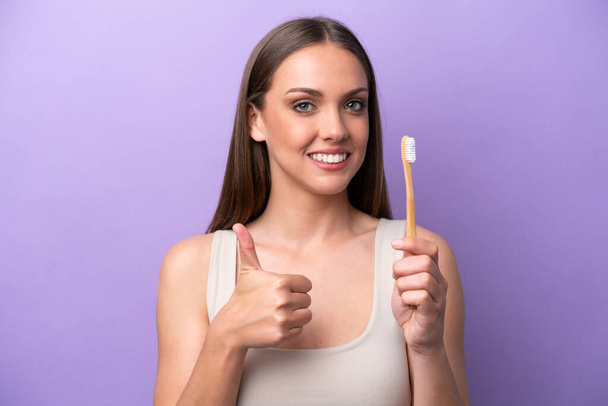 Young caucasian woman brushing teeth isolated on purple background with thumbs up because something good has happened - Foto, Imagen