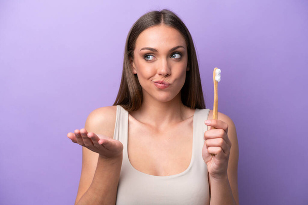 Young caucasian woman brushing teeth isolated on purple background making doubts gesture while lifting the shoulders - Foto, Imagen