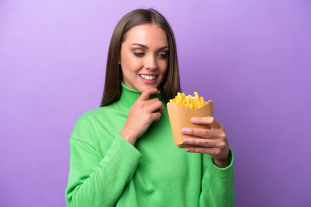 Молодая белая женщина с жареными чипсами на фиолетовом фоне смотрит в сторону и улыбается - Фото, изображение
