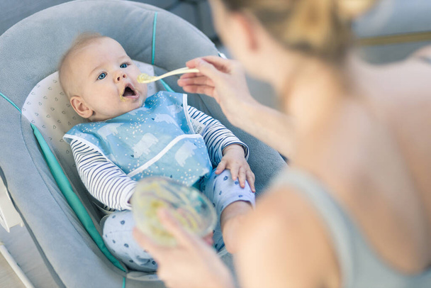 Mother spoon feeding her baby boy child in baby chair with fruit puree. Baby solid food introduction concept - Photo, Image
