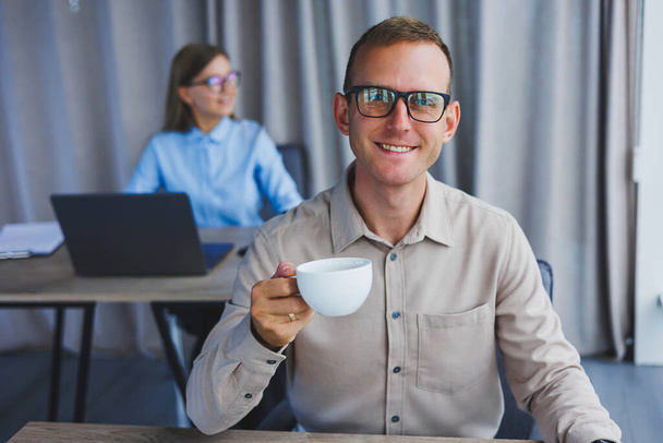 European businessman looking at something on laptop while working and drinking coffee. The concept of a modern successful person. Young guy in glasses sitting at the desk in the office with a laptop - Zdjęcie, obraz