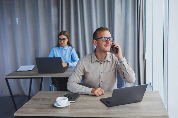 A handsome businessman is working during a video call on a laptop. The concept of a modern successful person. Young smiling male entrepreneur in glasses sits and works at a desk in an office space - Photo, Image